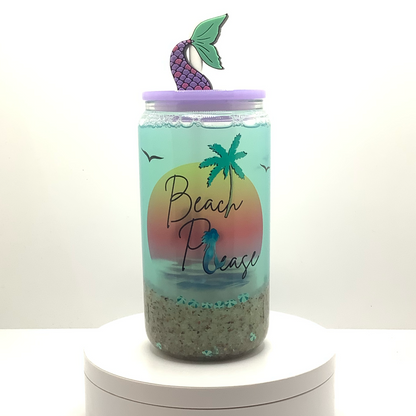 16ox glass can cup with 'Beach Please' text and palm tree, mermaid design sublimated on a clear snow globe tumbler. Snow globe wall is filled with sand, seashell polymers, and blue green water. Light purple lid and mermaid tail straw topper 