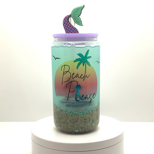 16ox glass can cup with 'Beach Please' text and palm tree, mermaid design sublimated on a clear snow globe tumbler. Snow globe wall is filled with sand, seashell polymers, and blue green water. Light purple lid and mermaid tail straw topper 